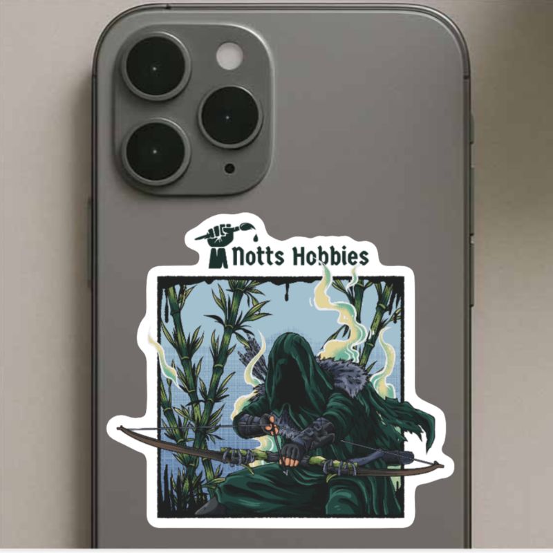 A die-cut vinyl sticker with a white border is shown applied to the back of a gray smartphone. The sticker features the text 'Notts Hobbies' above an illustration of a hooded figure in green robes holding a bow, set against a background of tall bamboo with yellow-green mist.